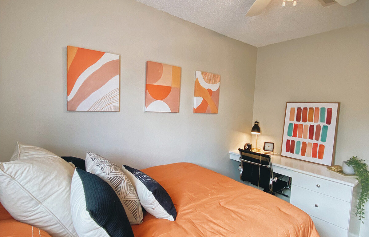 Model unit with a orange bed, desk with a table, chair, plants and artwork on the wall