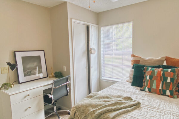 Model unit with bed with throw pillows and blanket. Nearby a overhead fan, closet, study desk with a light and leaning artwork, chair and exterior window
